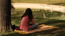 person with long dark hair sitting on a blanket under a tree gazing at a pond